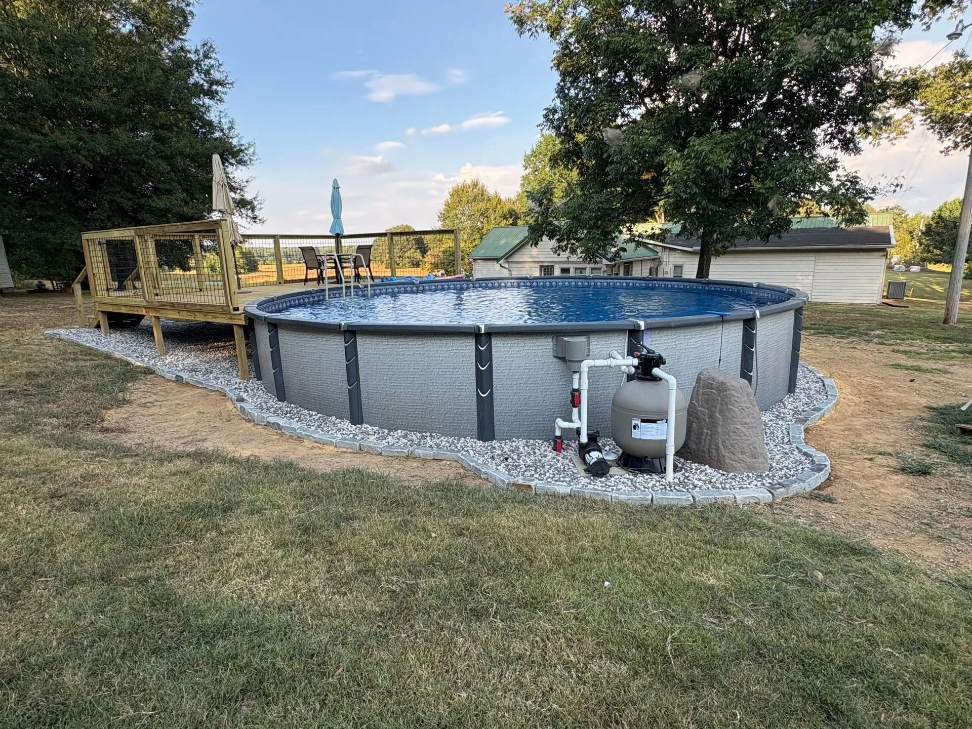 Above-ground pool in a grassy yard, surrounded by gravel. Filter and small deck visible.