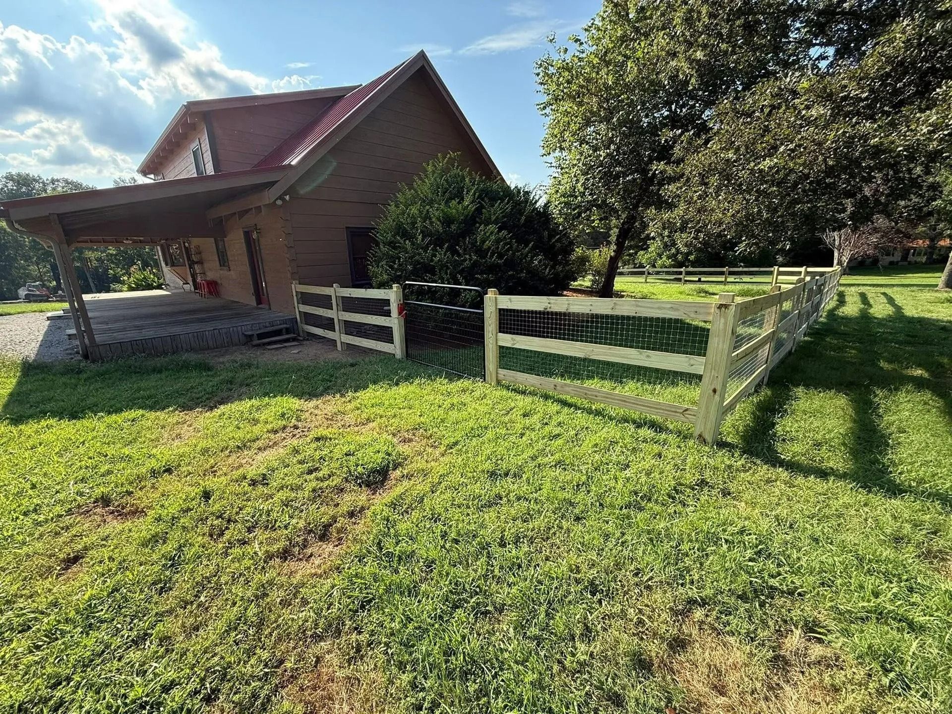 A wooden fence surrounds a grassy yard, leading to a house with a covered porch and red roof.