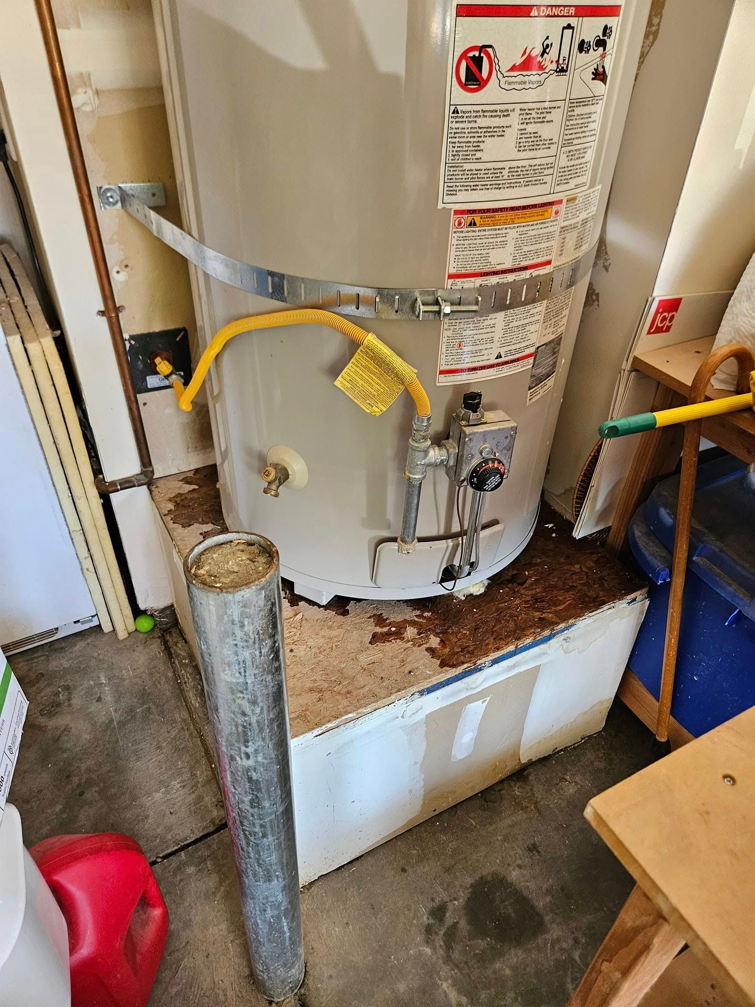 A water heater leaking onto a white, box-shaped platform in a cluttered garage setting.