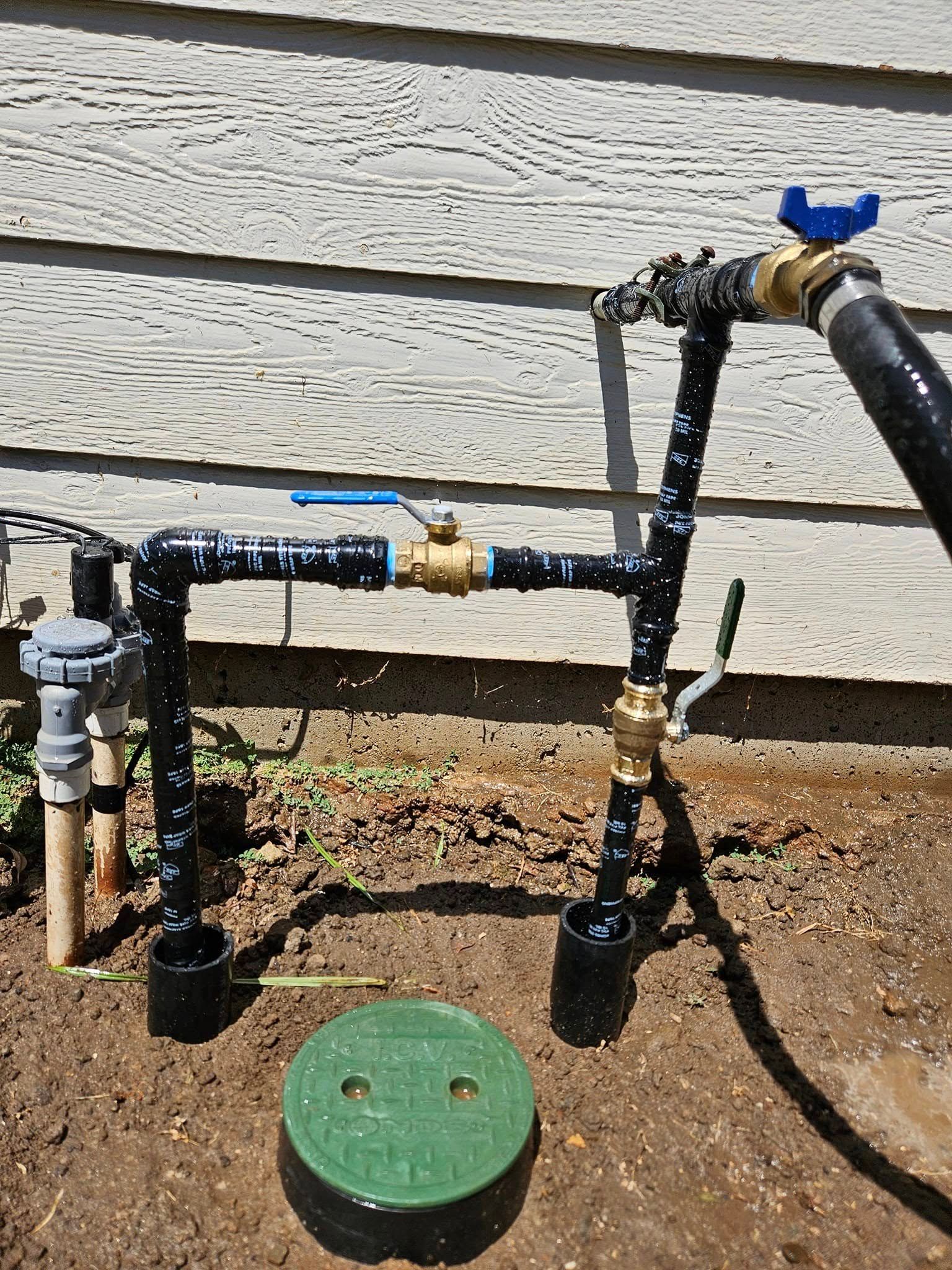 Water pipes and valves for irrigation system near a house exterior. Green cap in the foreground.