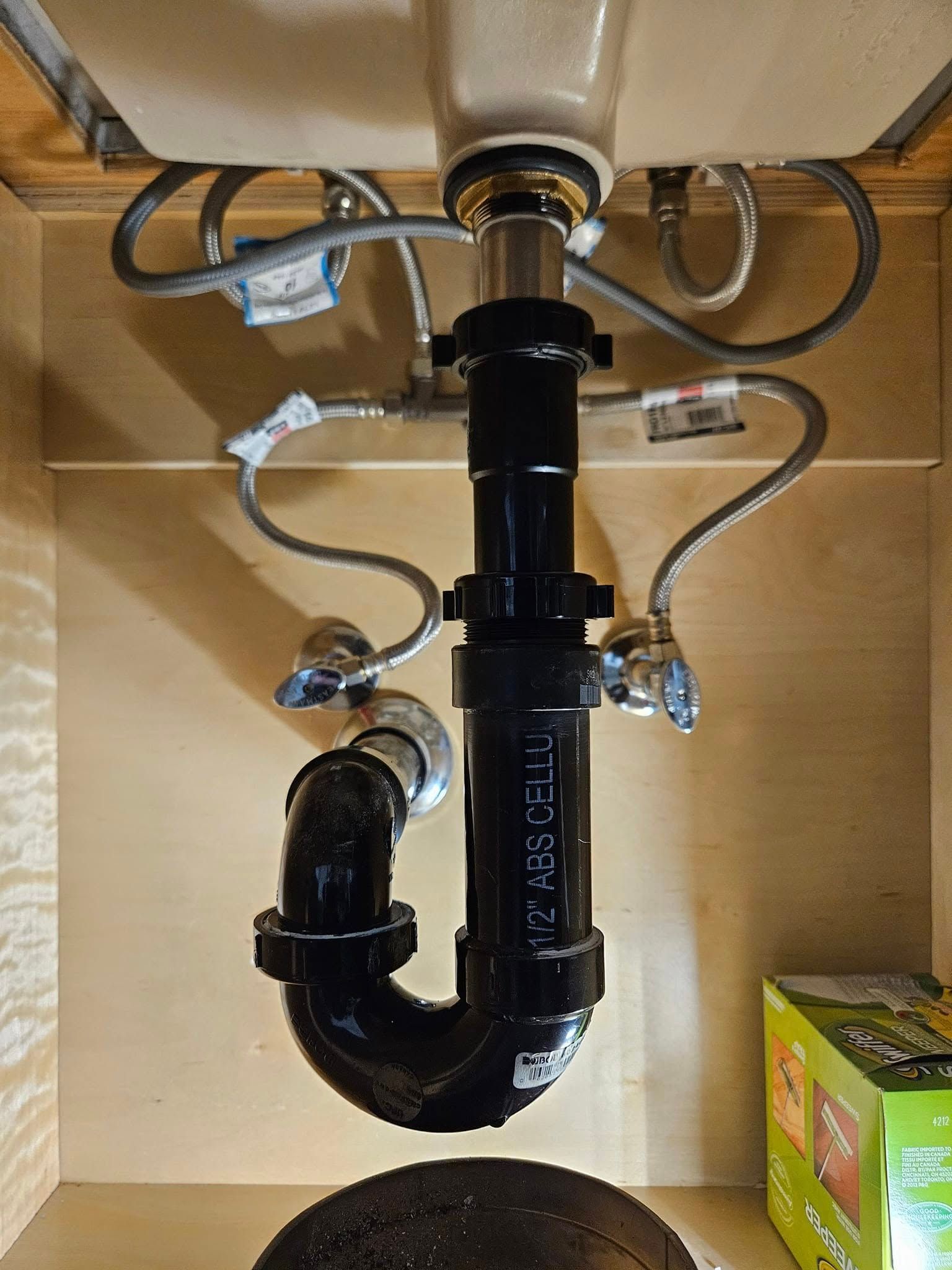 Underside of a bathroom sink showing plumbing: black P-trap, drain pipes, and water supply lines against a wooden cabinet.