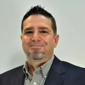 Man in a navy suit and patterned shirt, with a goatee, smiling in front of a white background.