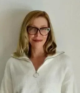 Woman with blonde hair, glasses, wearing a white sweater, smiles at the camera.