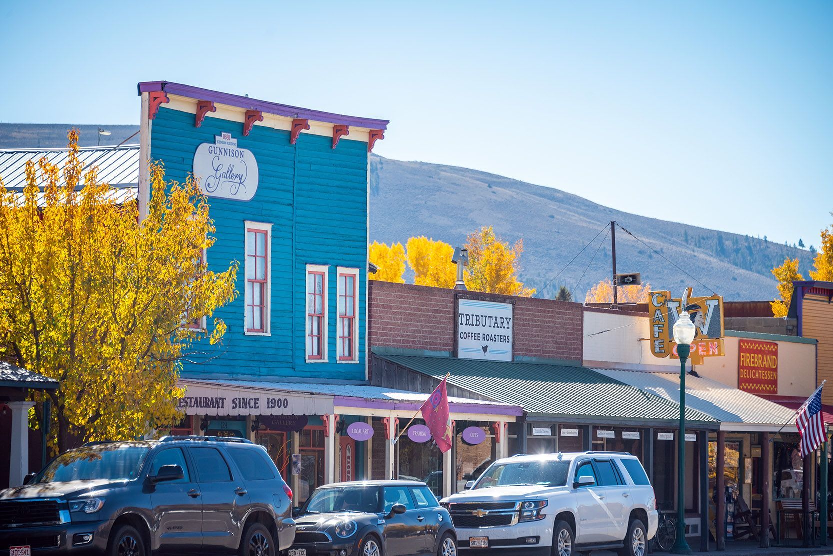 Colorful buildings line a street with parked cars and fall foliage. Mountains in the background.