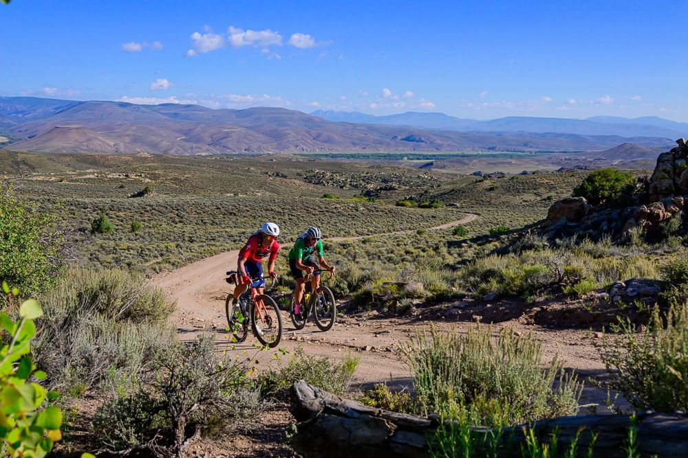 Two cyclists riding mountain bikes on a dirt path in a desert landscape under a blue sky.