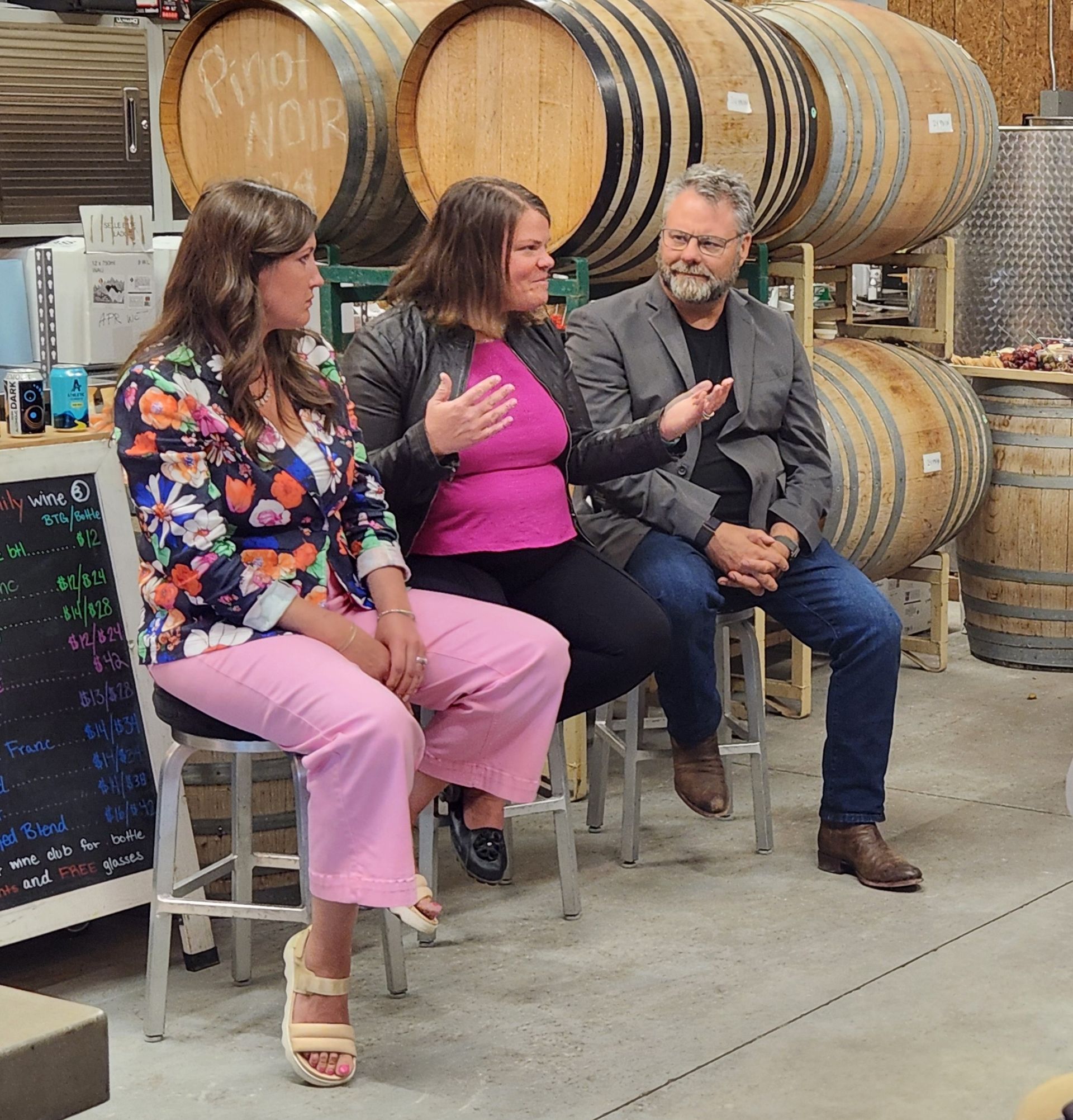 Three people sitting and talking in a winery. A woman in pink gestures, others listen. Barrels in the background.
