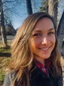 Woman smiling outdoors with long brown hair, wearing a dark jacket over a red plaid shirt, trees in background.