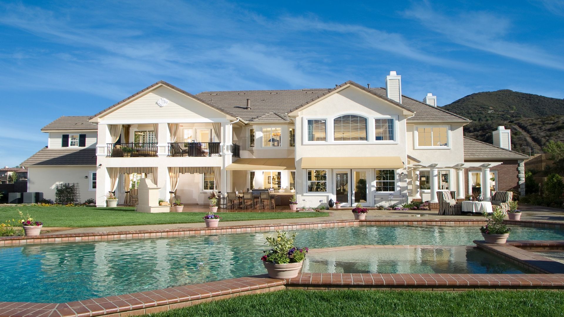 Luxury home exterior with a swimming pool, patio, and mountain backdrop under a blue sky.