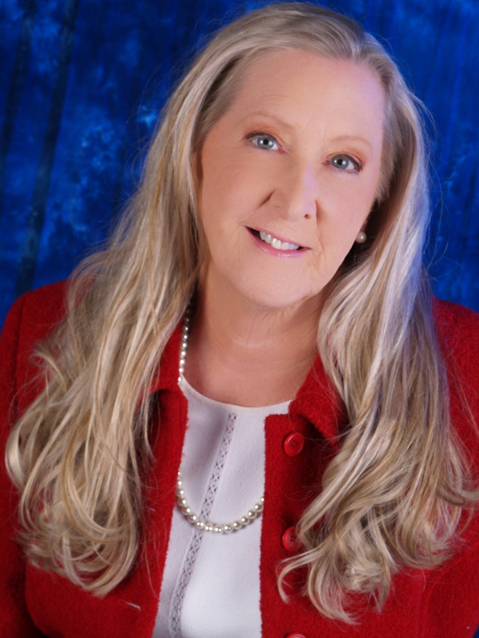 Blond woman with long hair wearing a red jacket and pearl necklace, smiling against a blue background.