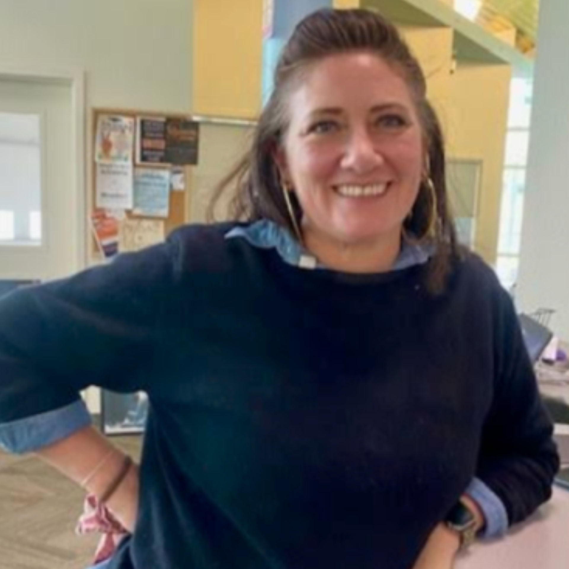 Woman smiling, wearing a blue sweater over a denim shirt, hoop earrings, indoors.