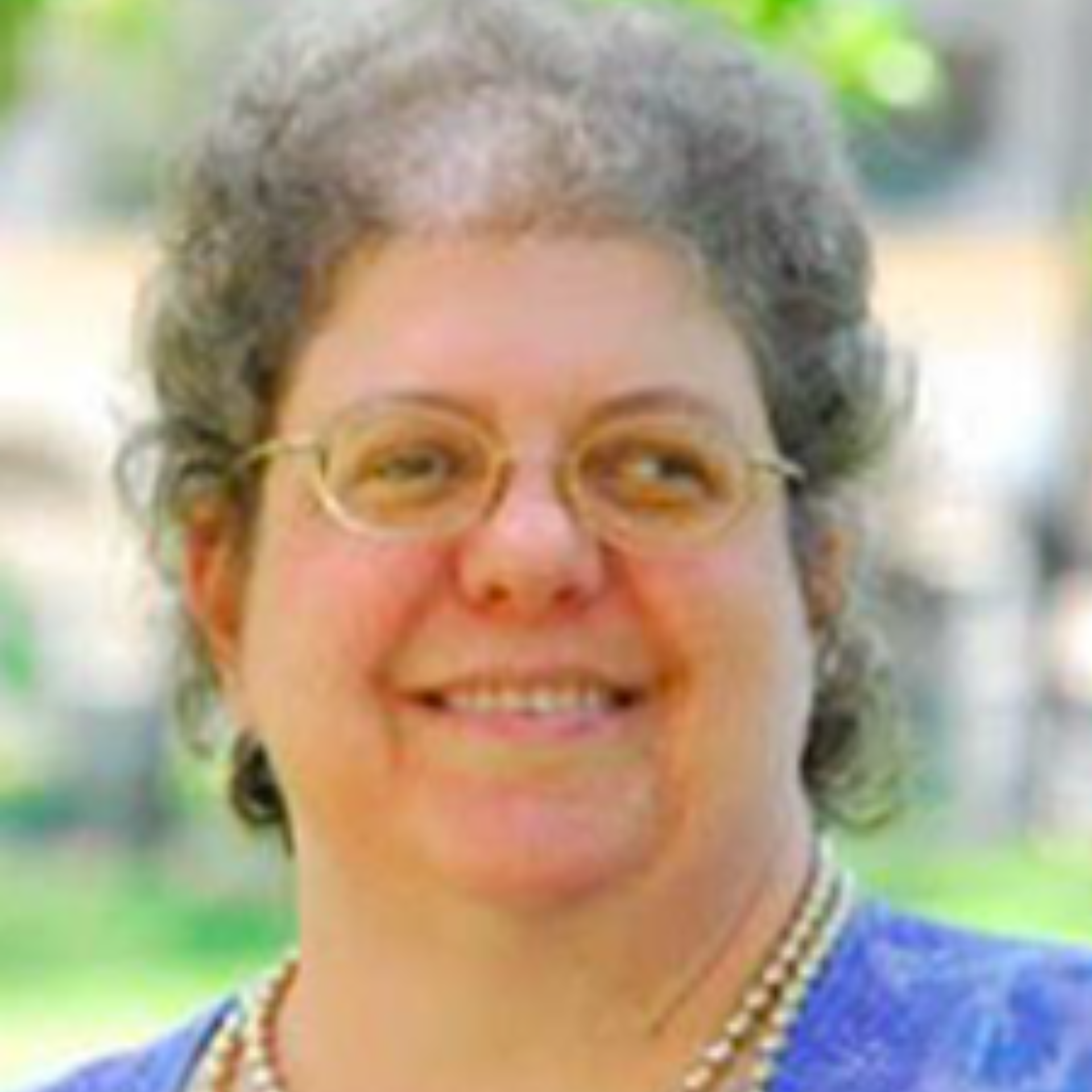 Woman with glasses smiling, wearing a blue shirt and pearl necklace, outdoors.