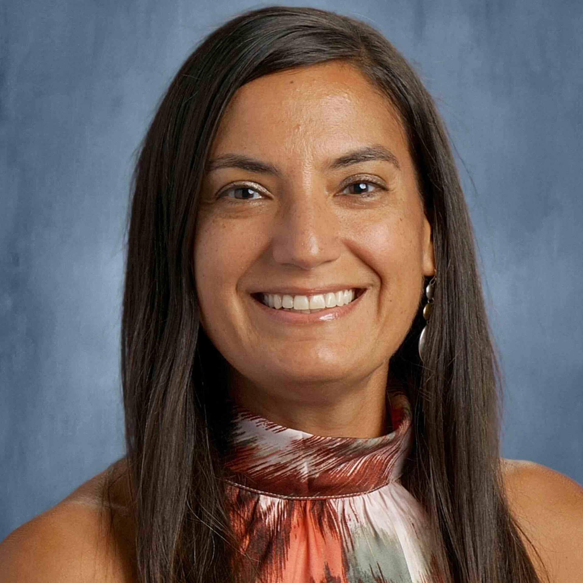 Woman with brown hair smiling at the camera, wearing a patterned top, blue background.