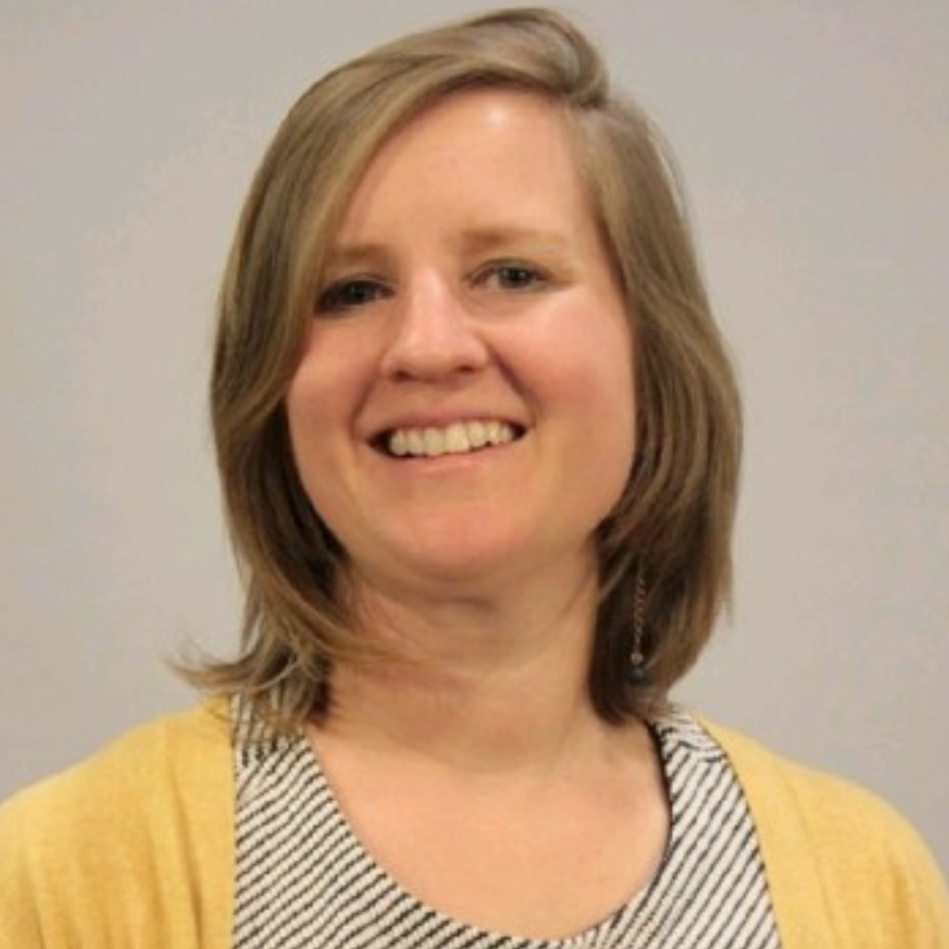 Woman smiles, shoulder-length blonde hair, wearing a striped shirt and yellow cardigan. Background: neutral wall.