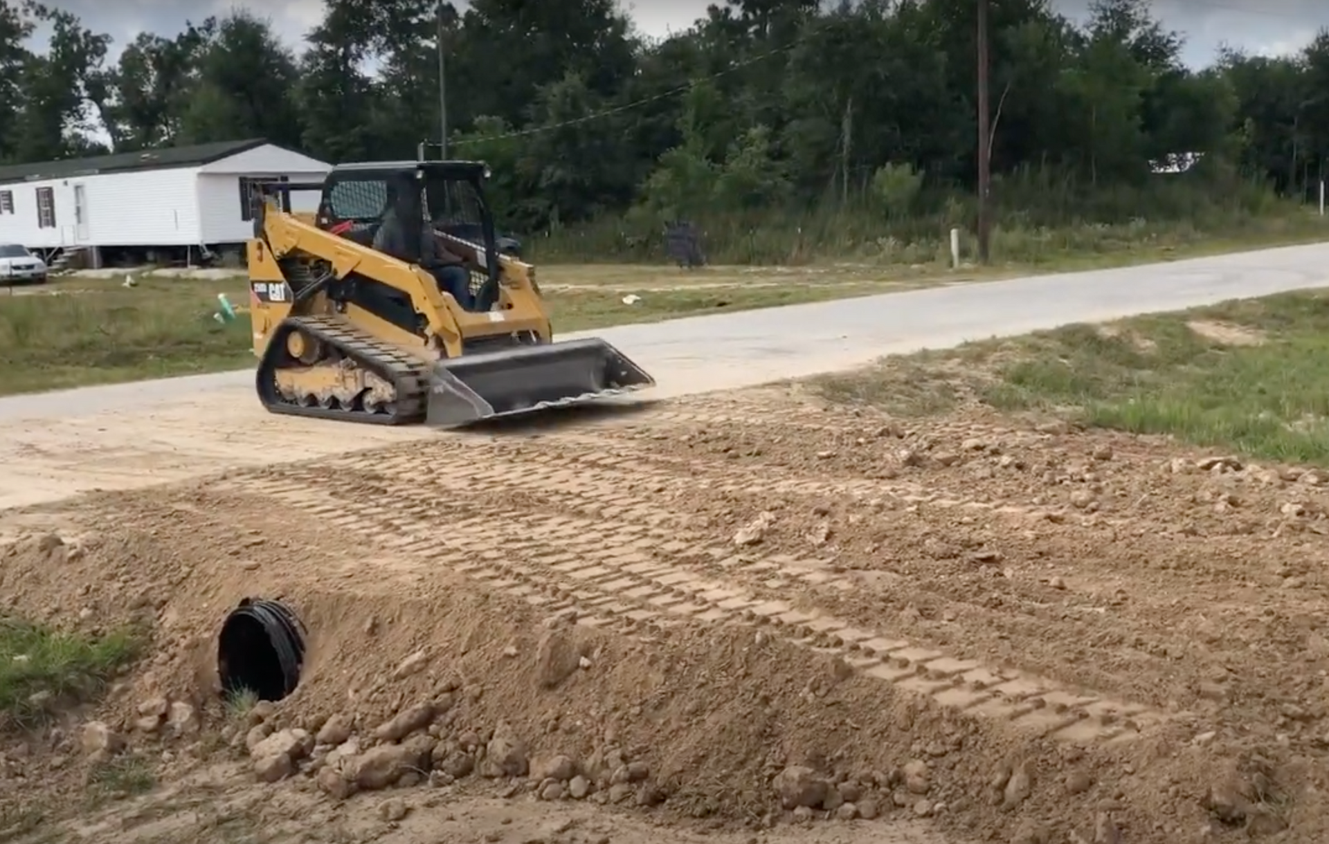 Trackloader grading over the top of a freshly installed culvert in front of a white mobile home in rural Texas.