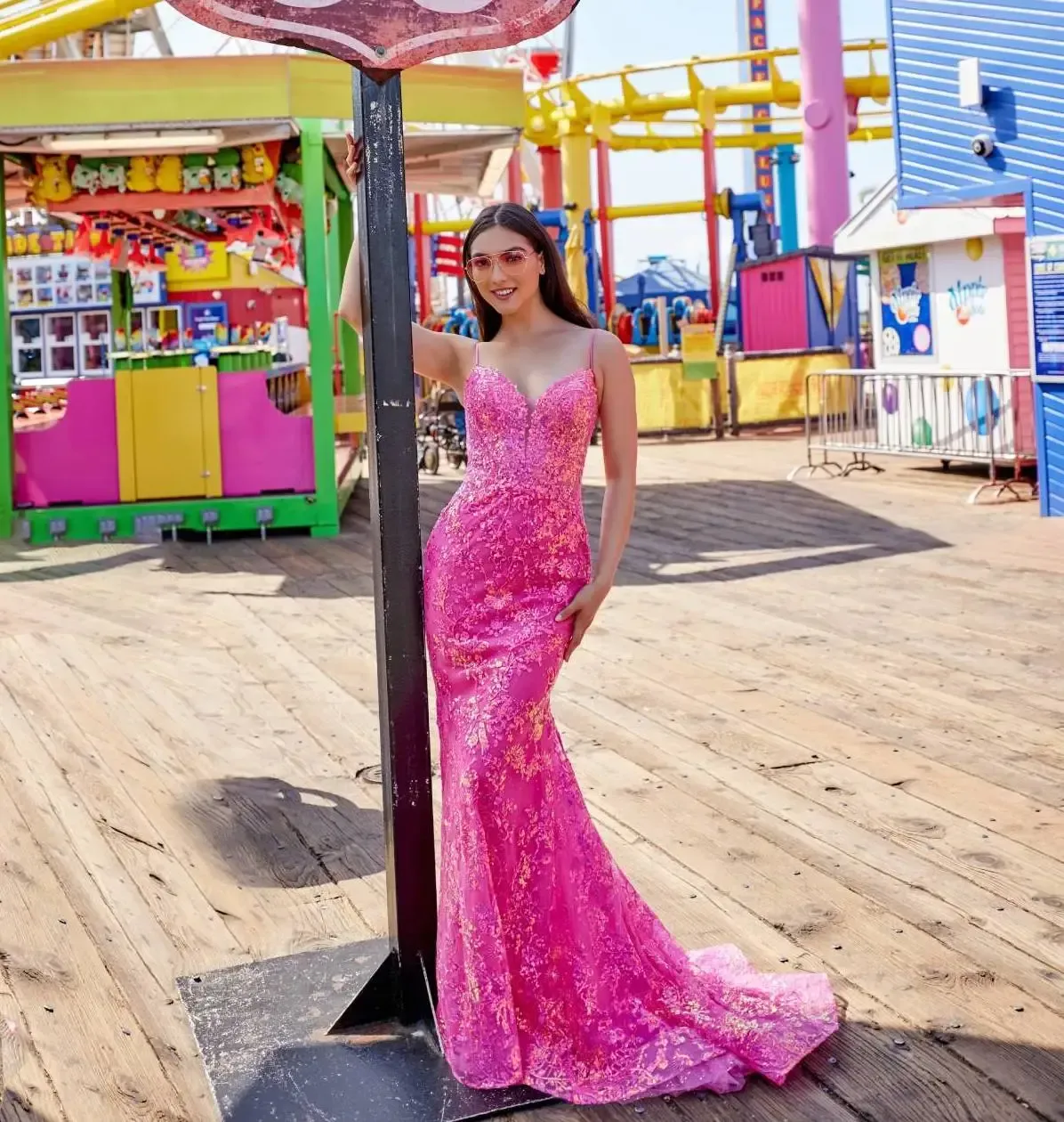 Woman in a sparkly pink gown poses next to a sign on a boardwalk with colorful amusement park attractions.