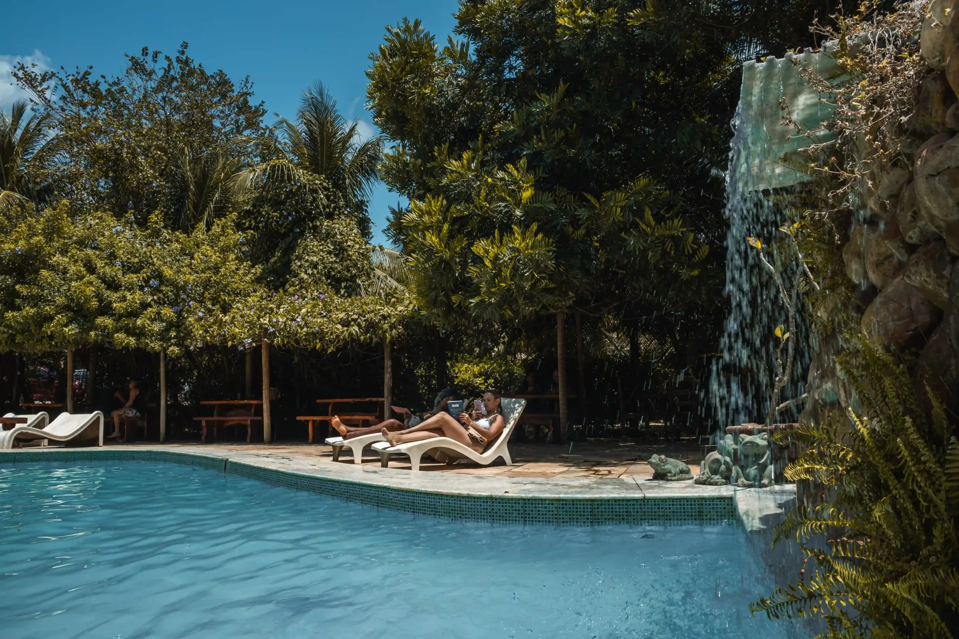 Algumas pessoas estão sentadas em espreguiçadeiras ao lado de uma piscina com uma cachoeira ao fundo.-Pousada-Imperio-do-Sol