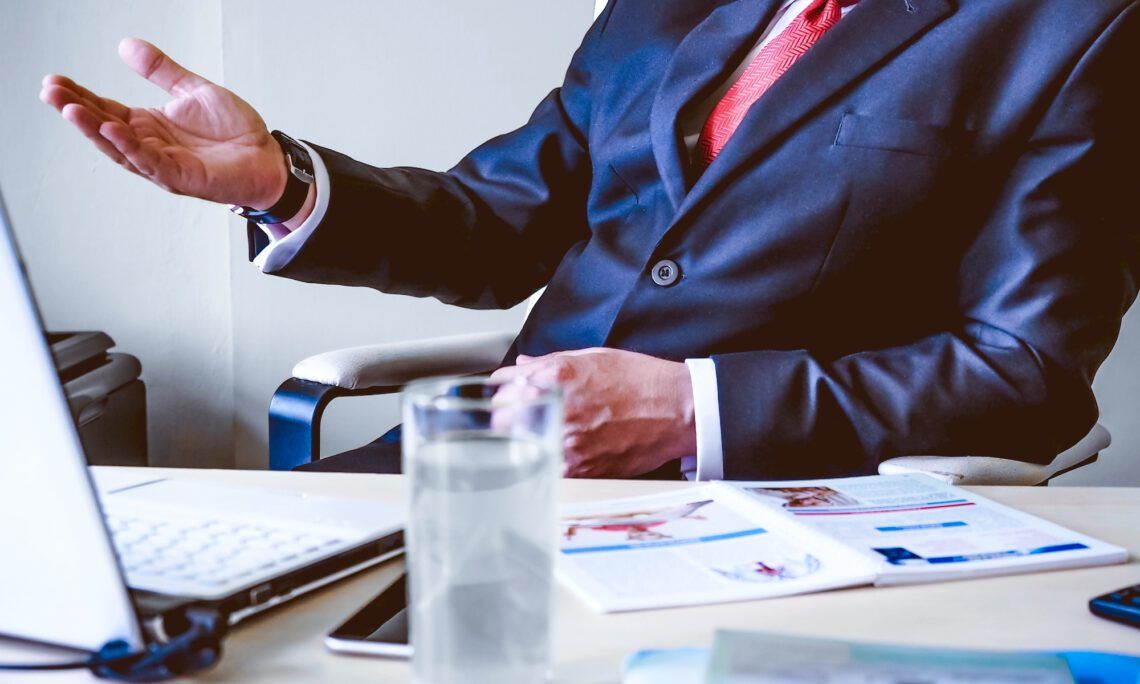 A man in a suit and tie is sitting at a desk with a laptop and a glass of water.