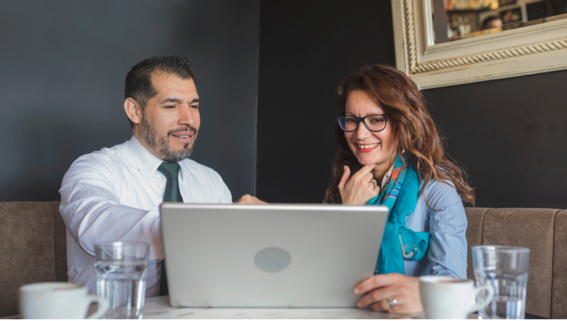 A man and a woman are sitting at a table looking at a laptop computer.