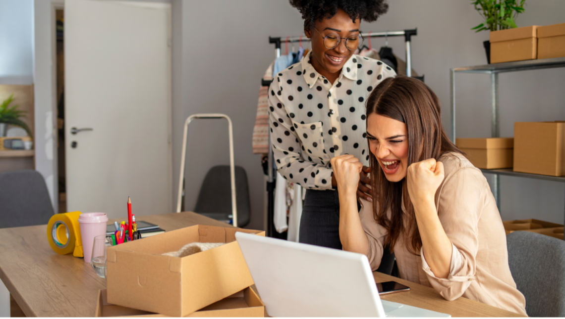 Two women are sitting at a desk looking at a laptop computer.