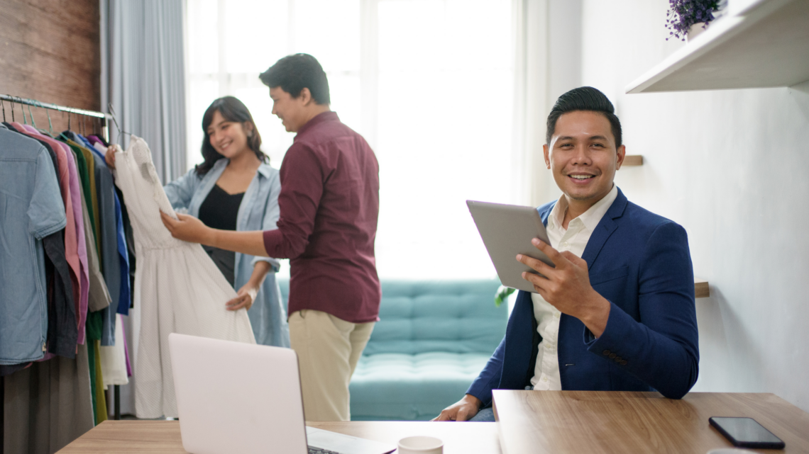 A man is sitting at a desk holding a tablet and a woman is holding a dress.