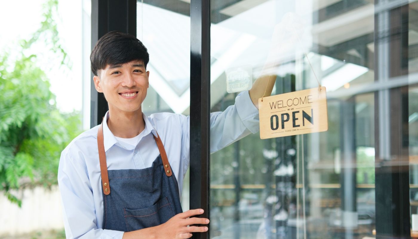 Young male business owner holding a store sign in front of his shop, showcasing the impact of franchise consultant services on new franchise owners.