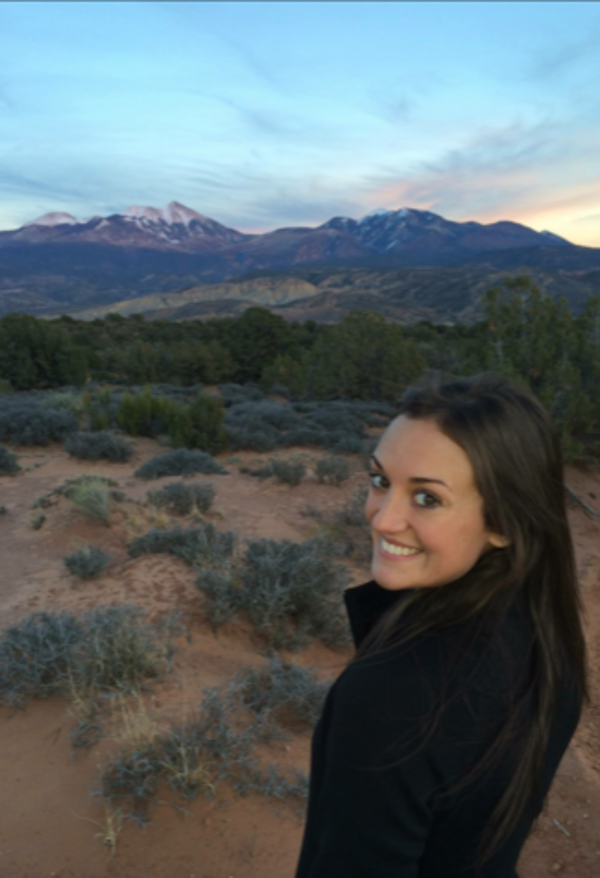 Woman in sunglasses and off-the-shoulder dress smiles on a beach with turquoise water.