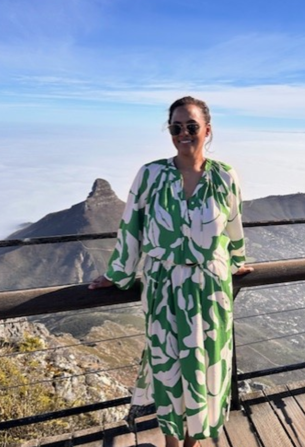 Woman in sunglasses and off-the-shoulder dress smiles on a beach with turquoise water.