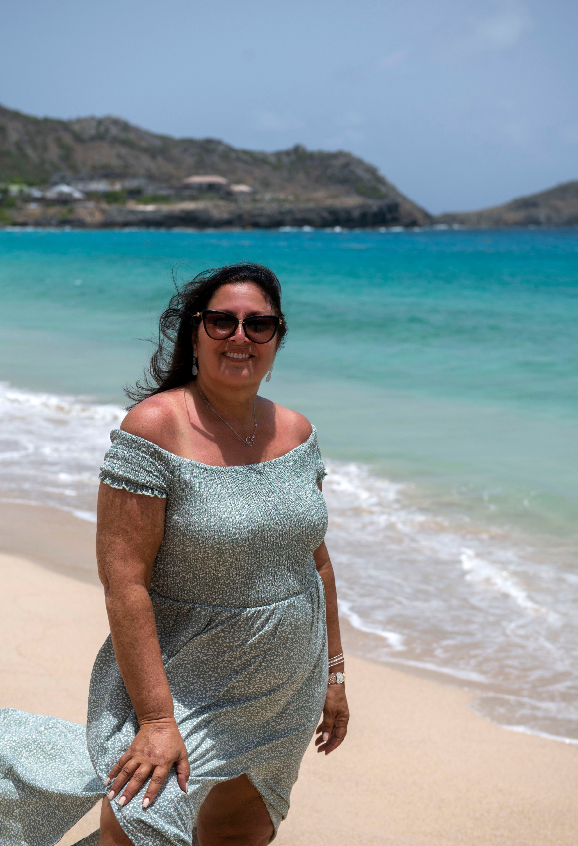 Woman wearing sunglasses and yellow top, smiling in front of a pink and white striped wall.