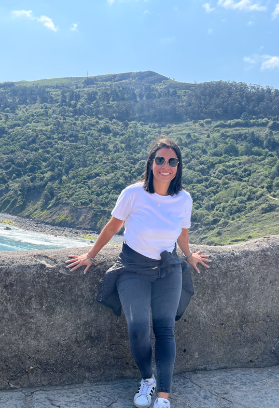 Woman in sunglasses and off-the-shoulder dress smiles on a beach with turquoise water.
