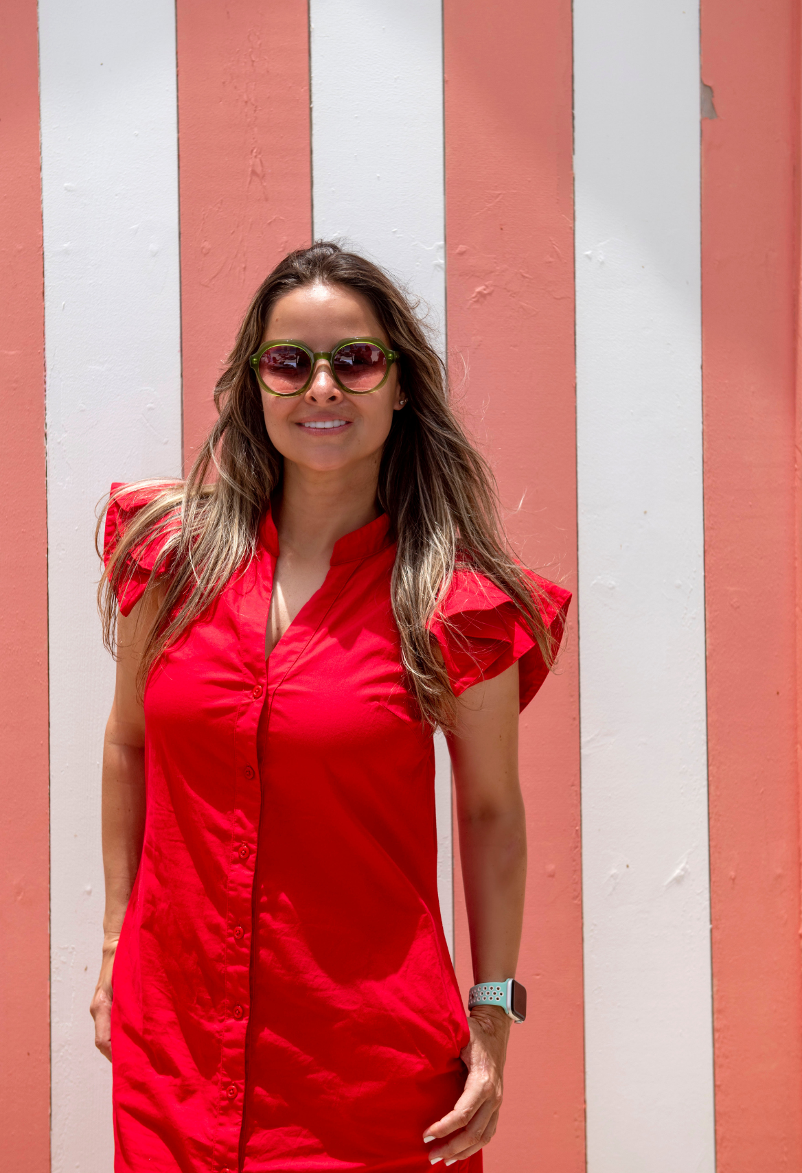 Woman in red dress and sunglasses, standing against a pink and white striped wall.
