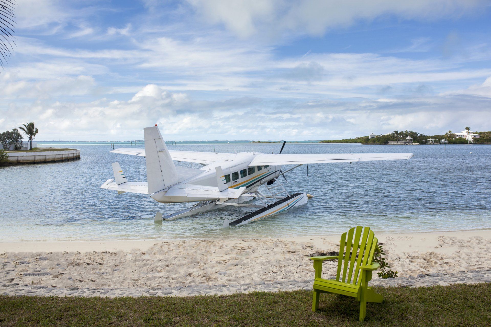 Seaplane on water near a sandy beach with a green chair. Bright blue sky and water.