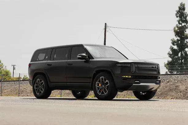 Black Rivian SUV parked on a road with a clear sky and industrial background.