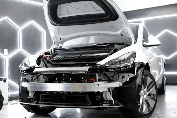 White Tesla Model Y with front bumper removed in repair shop, hood open.