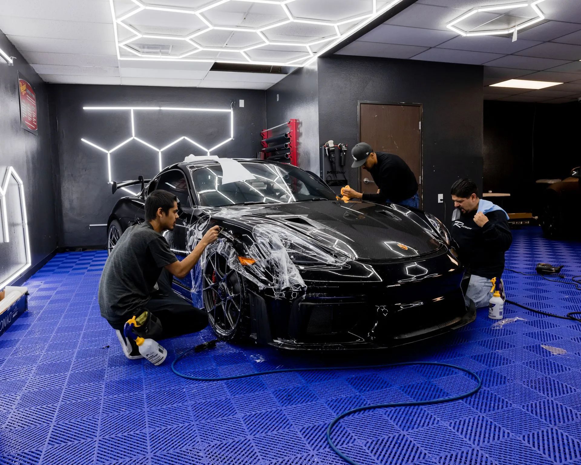 Three people washing a black car in a garage with blue tiled floor and hexagon lights.