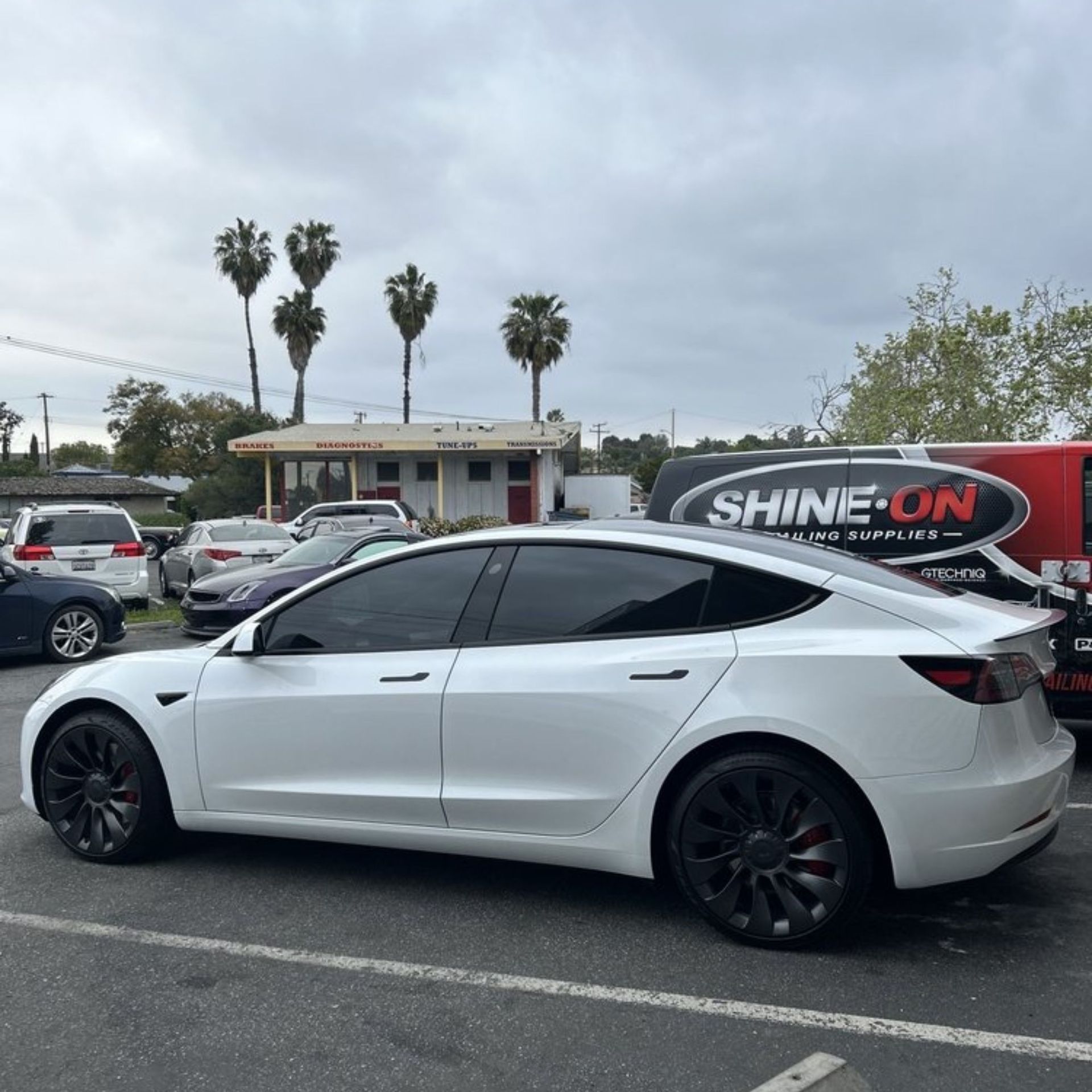 Hand wiping a shiny black car wheel, red car in the background.