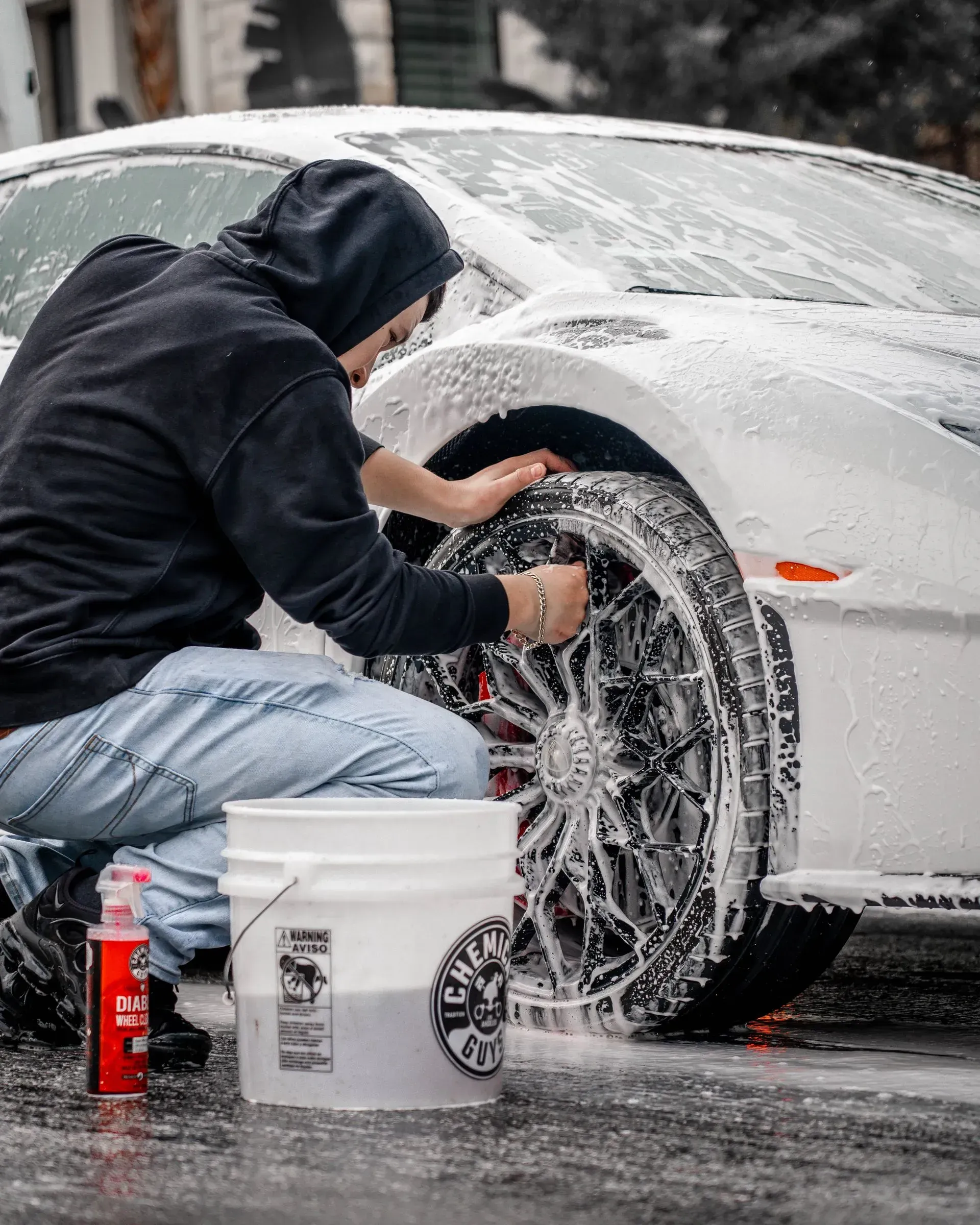 Person washing a white car wheel with a bucket and soap in an outdoor setting.