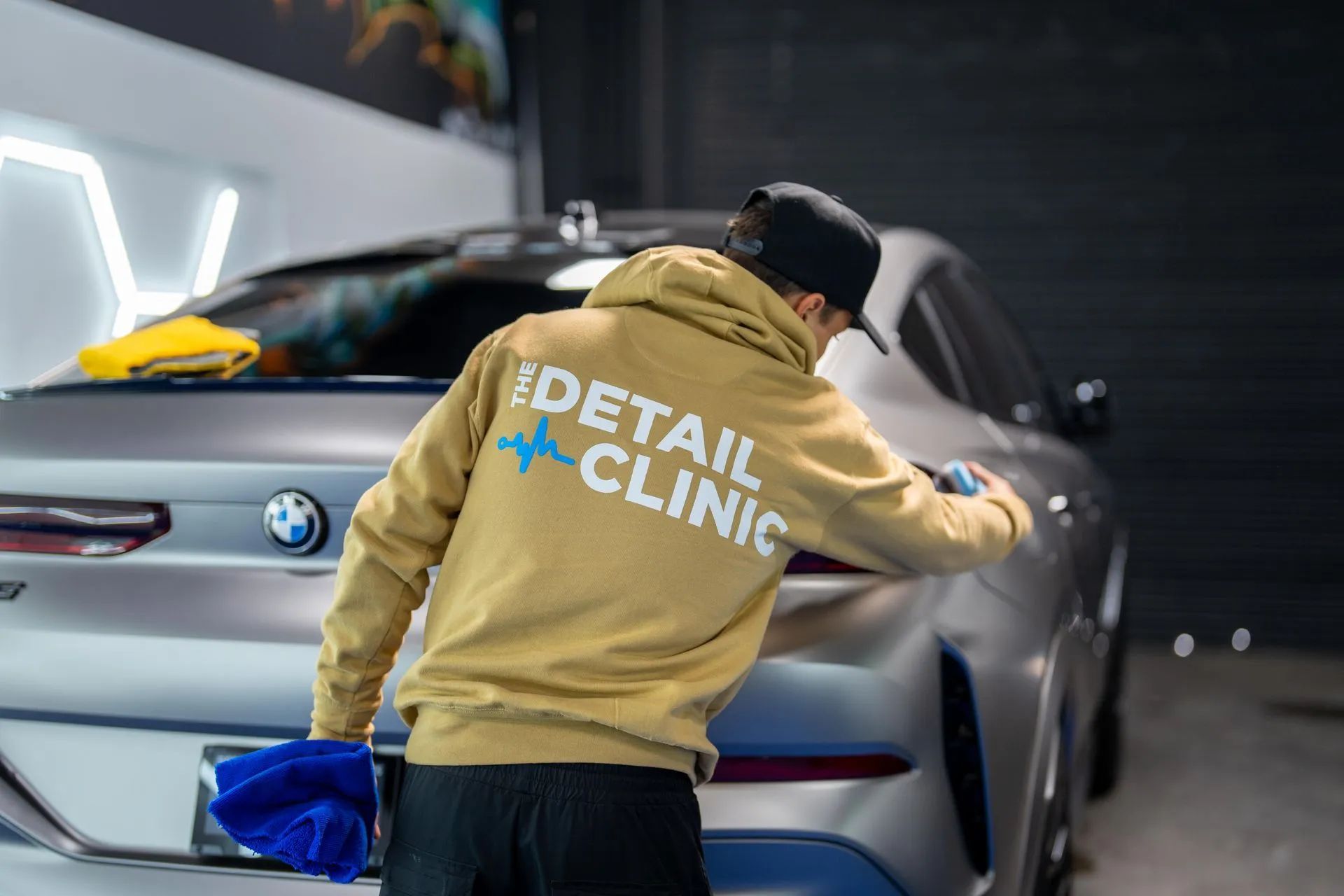 Person wearing tan hoodie polishing a silver BMW car at a detail clinic.