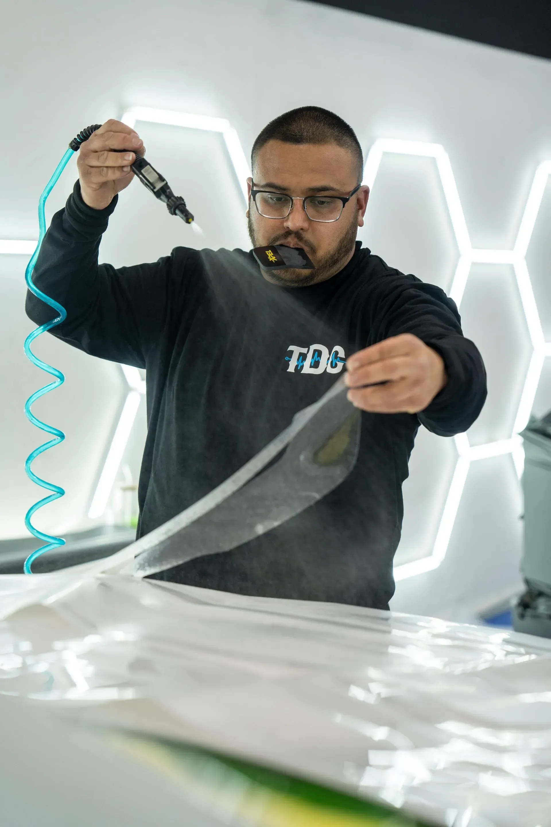 Man applying a protective film to a car, using a heat gun. Indoors, against a white surface.
