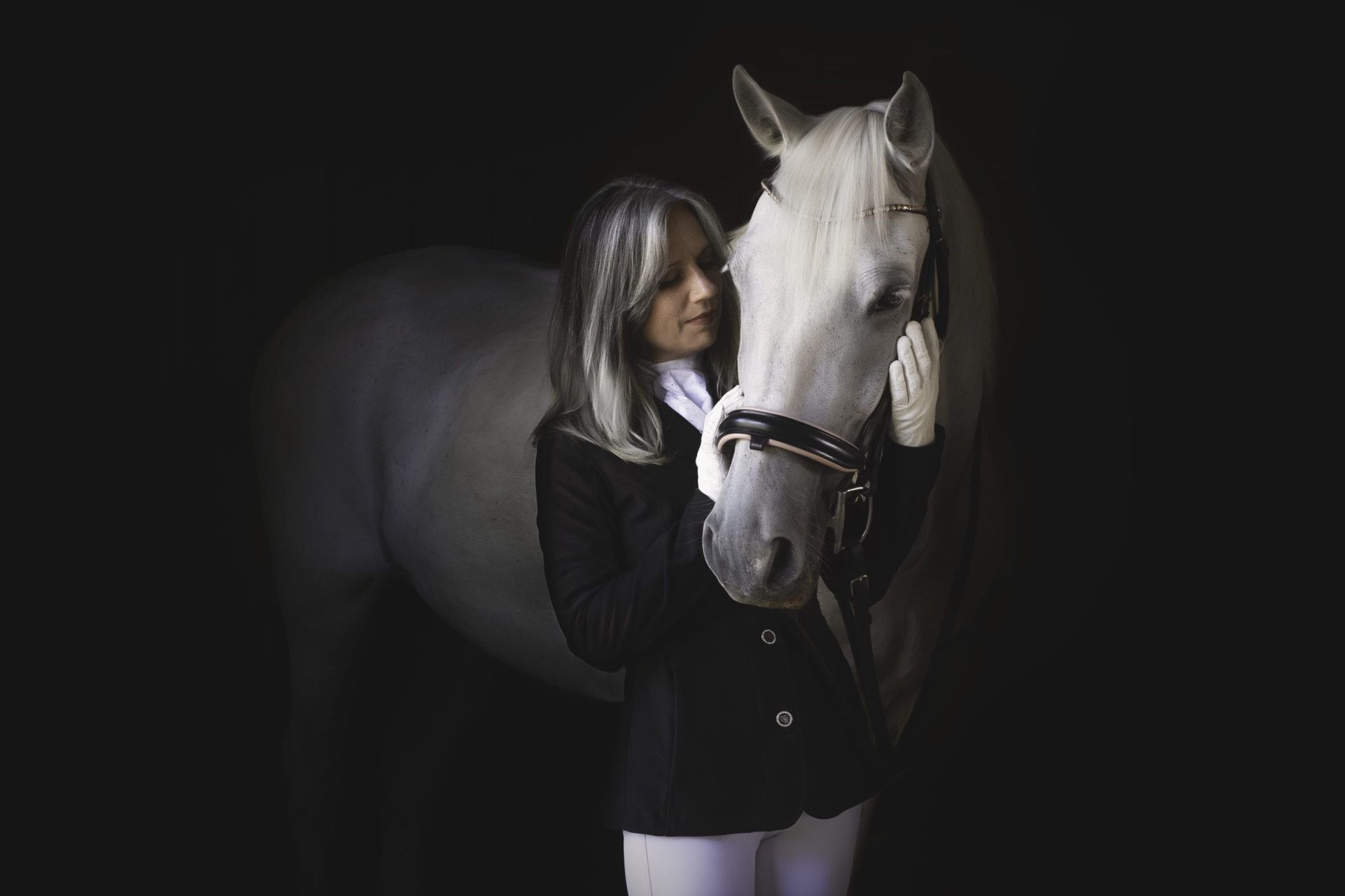 Woman in riding attire, embraces a white horse, black backdrop.