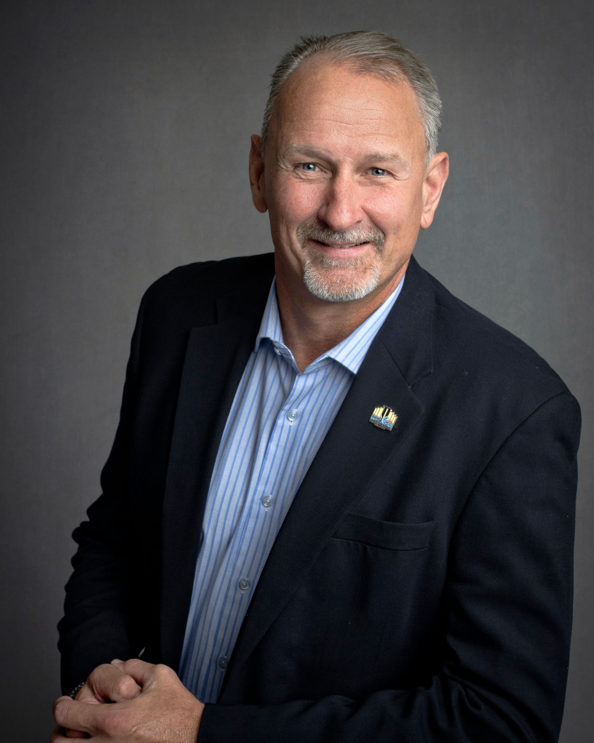 Man in blue shirt and blazer, smiling, hands clasped, set against gray background.