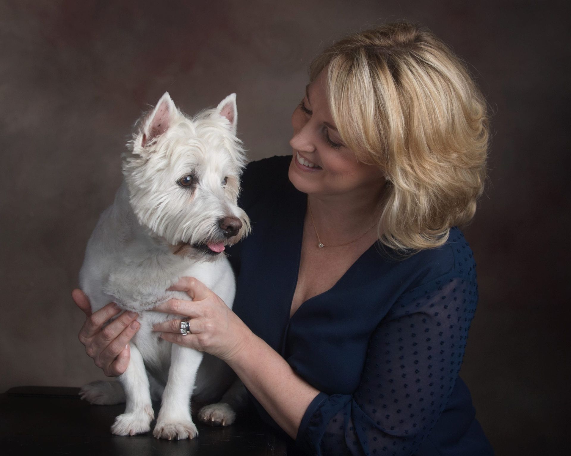 Woman with blonde hair smiles at a white West Highland Terrier, both on a brown background.