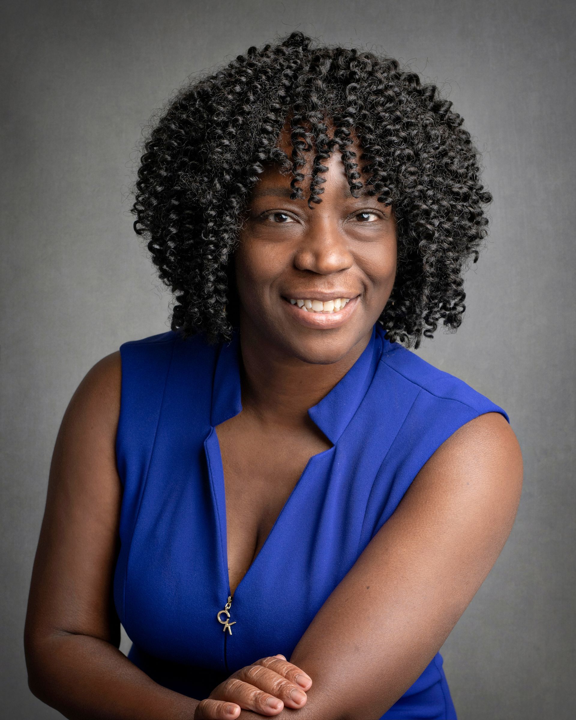 Woman with dark skin and curly hair in a blue dress smiles against a gray backdrop.