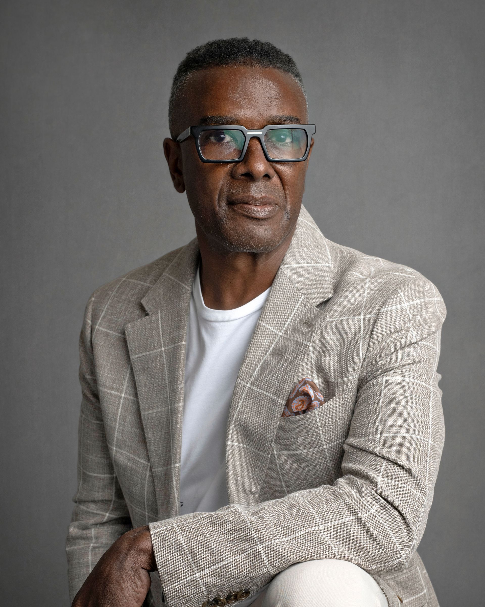 Older Black man with glasses, in blazer and white shirt, looking forward against a gray background.