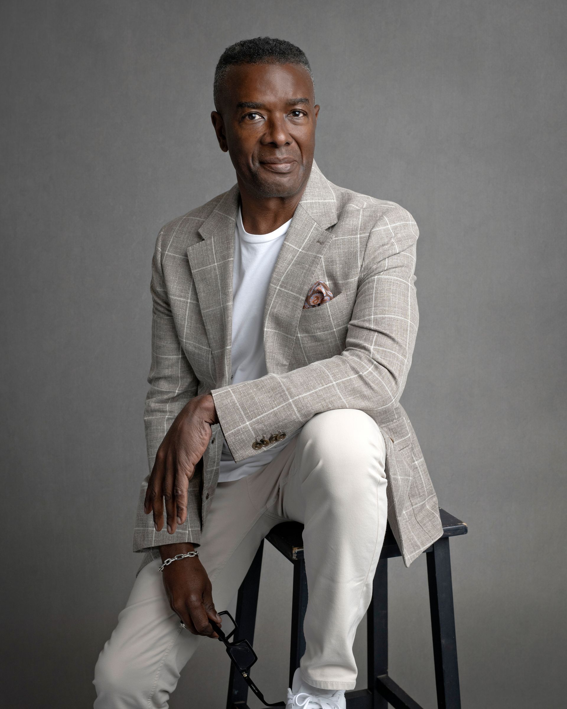 Mature Black man in blazer and slacks, seated on a stool, holding glasses.