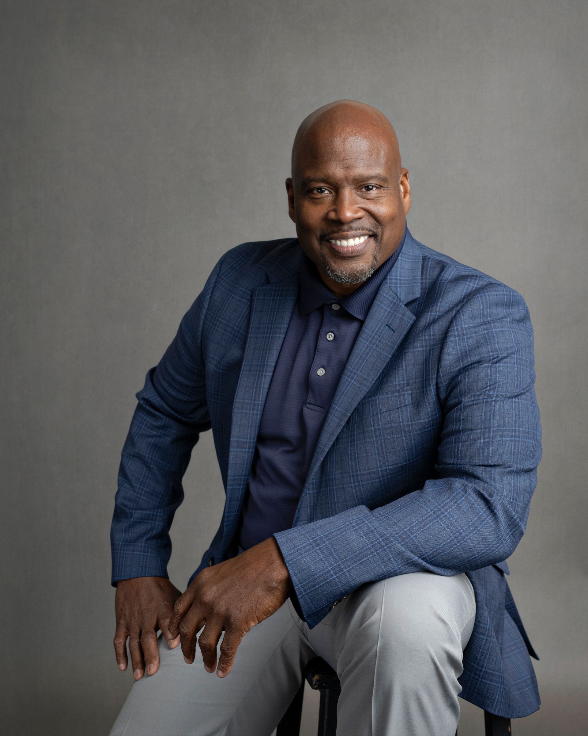 A smiling Black man in a blue blazer and polo, sitting on a stool, gray background.