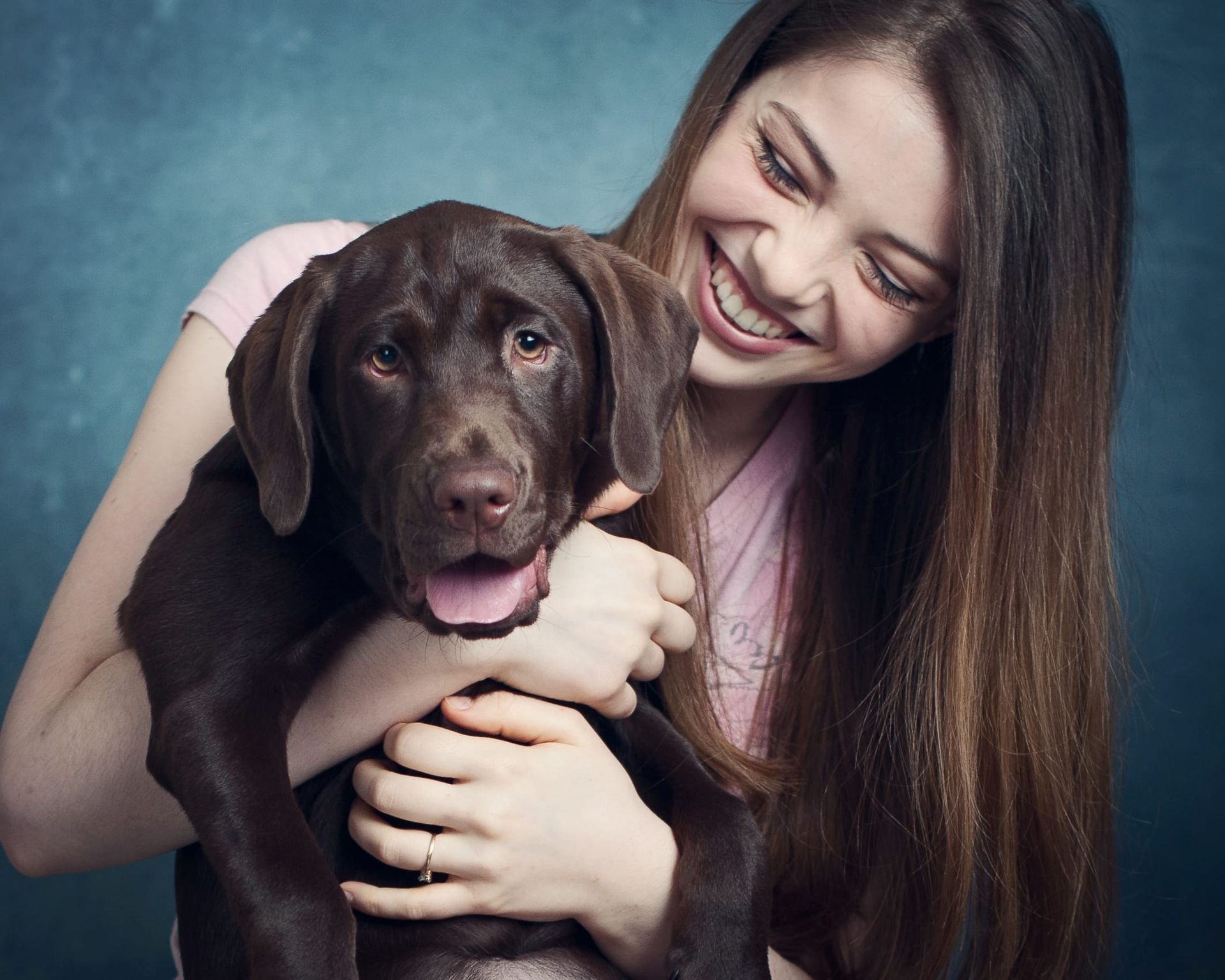 Woman with long brown hair happily hugging a chocolate Labrador puppy against a teal background.