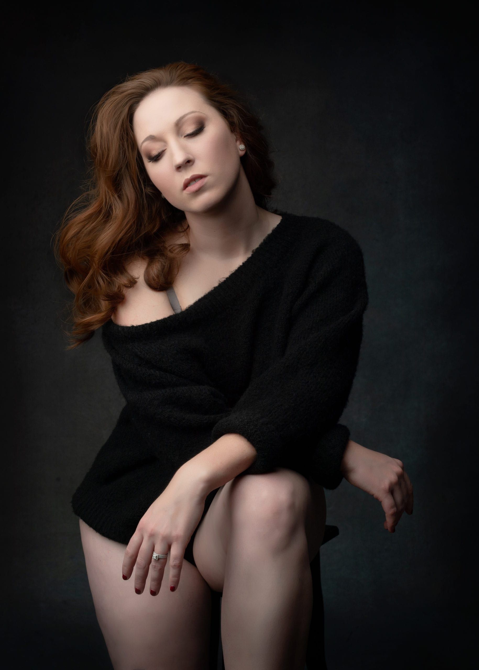 Woman with red hair, wearing a black sweater, sitting with eyes closed. Dark background.