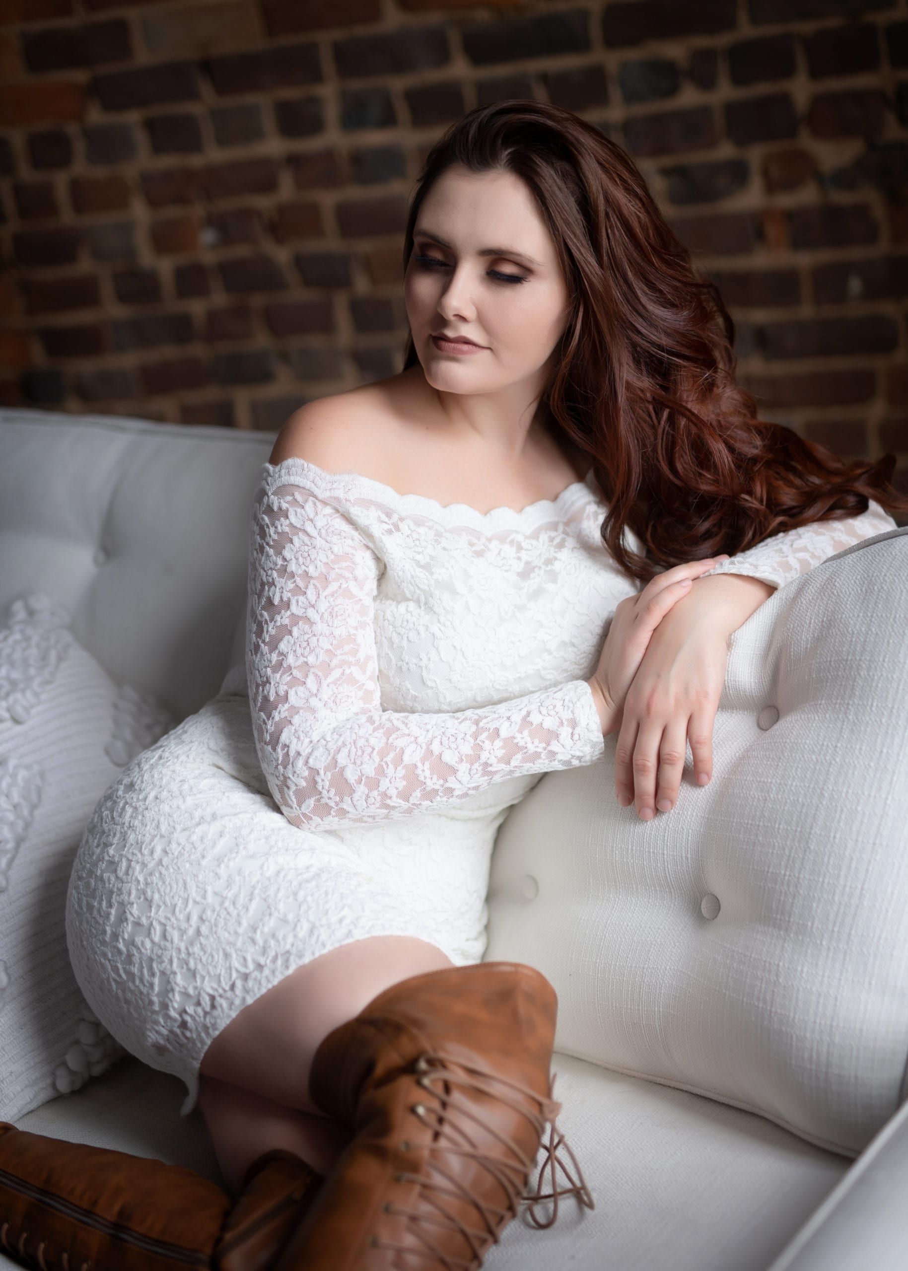 Woman in white lace dress and brown boots sits on white couch, red hair, brick wall background.