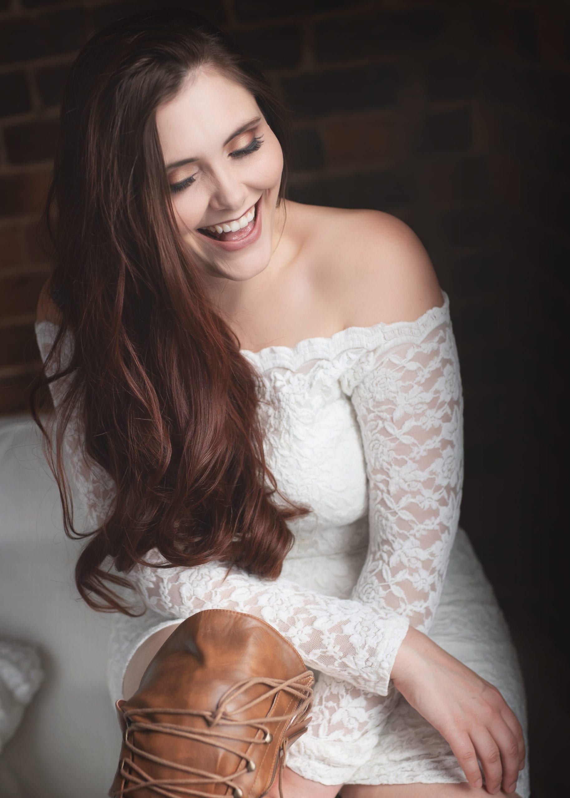 Woman in white lace dress laughs, leaning on brown boot in front of brick wall.