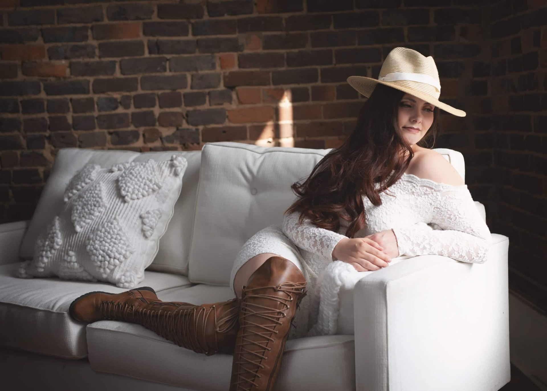 Woman in a white dress and cowboy boots, wearing a hat, reclines on a white couch against a brick wall.